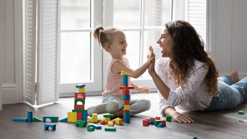 Happy mother playing wooden blocks with daughter, finish build tower, praising kid, giving high five settled down together on warm floor in living room. Playtime, help, happy motherhood, leisure games